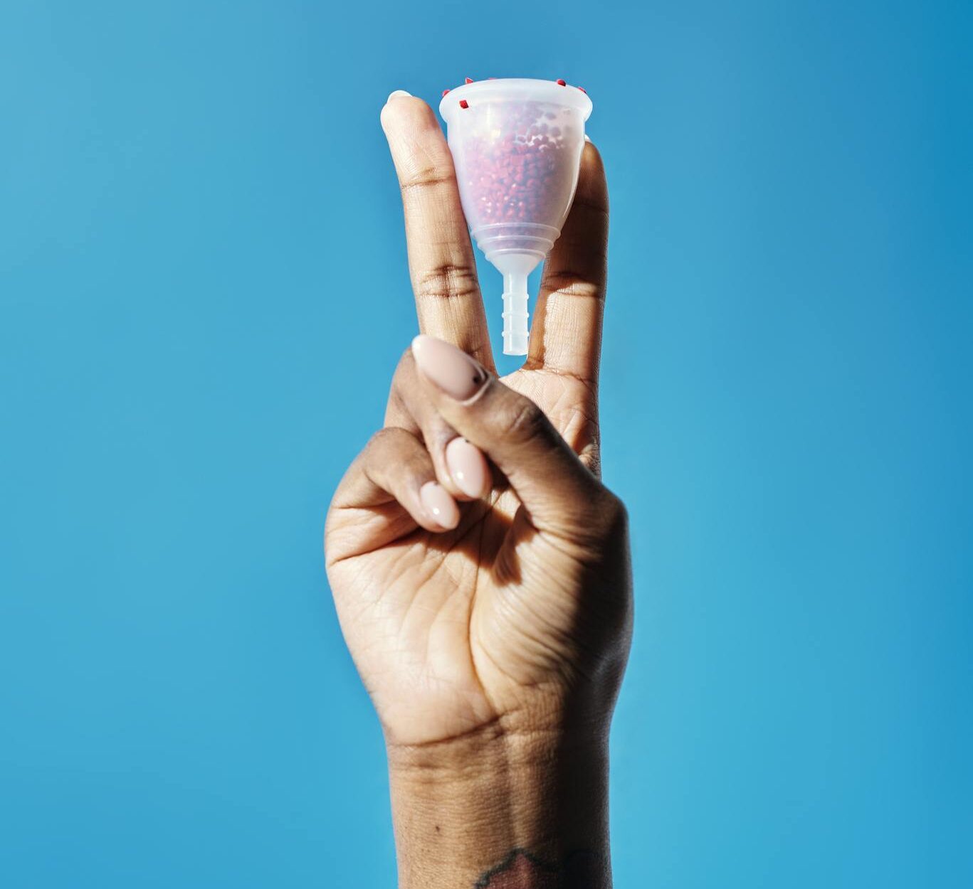 Close-up of a tattooed hand holding a menstrual cup in front of a bright blue background.